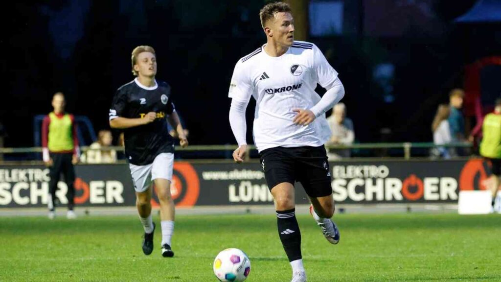 Ein Fußballspieler im weißen Trikot dribbelt den Ball auf dem Spielfeld, während ein Gegner im schwarzen Trikot verfolgt.