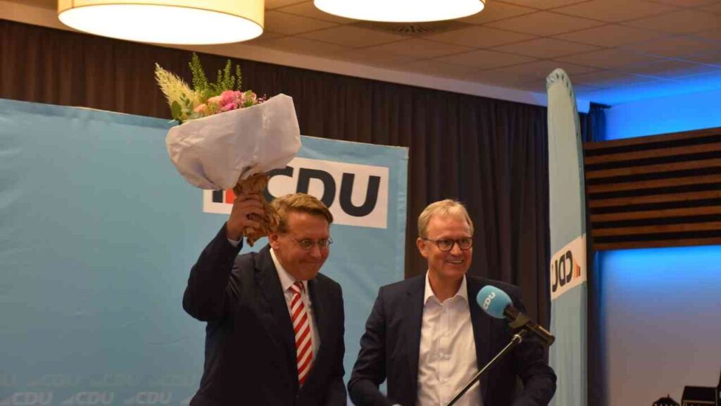 Politiker mit Blumenstrauß neben einem CDU-Rednerpodium bei einer Veranstaltung in einem Saal.
