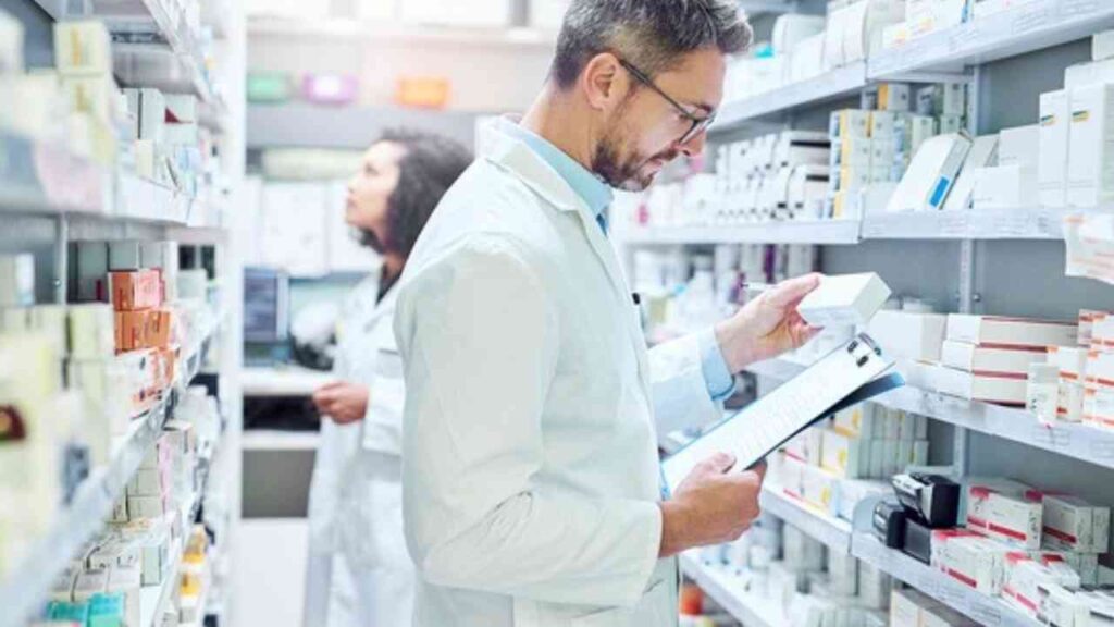 Apotheker überprüft Medikamente im Regal einer Apotheke, während Kollegin im Hintergrund arbeitet. Gesundheitswesen, Pharmazie.