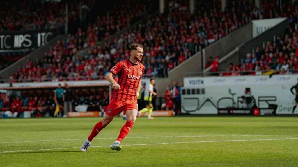 Fußballspieler in rotem Trikot während eines Spiels vor vollbesetzten Tribünen im Stadion.