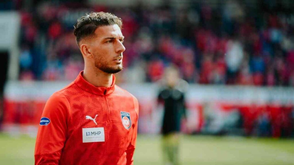 Ein Fußballspieler im roten Trikot steht auf dem Spielfeld, fokussiert auf das Spielgeschehen.