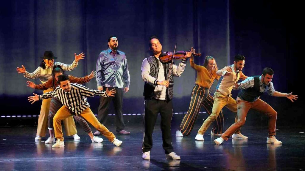 Tanzgruppe performt auf Bühne mit Geigenspieler im Mittelpunkt, dramatische Pose, bunte Kleidung und Scheinwerferlicht.