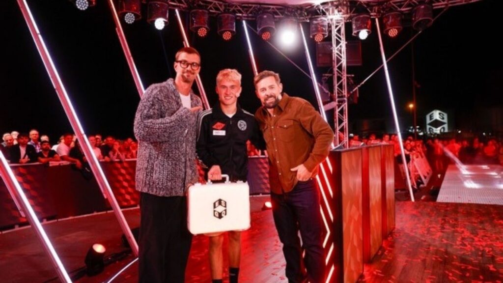 Drei Männer posieren bei einer Veranstaltung auf einer Bühne mit einem Lichtkoffer und Publikum im Hintergrund.