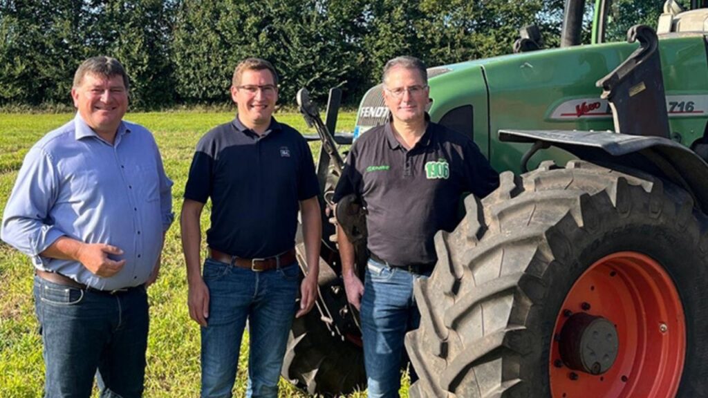 Drei Männer stehen neben einem grünen Traktor auf einem Feld, bei sonnigem Wetter.