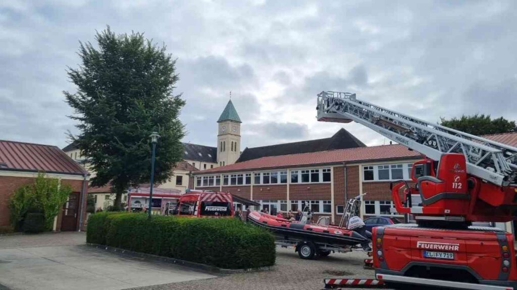 Feuerwehr im Einsatz vor einem Kirchengebäude mit Leiter und Boot auf Anhänger. Bewölkter Himmel im Hintergrund.