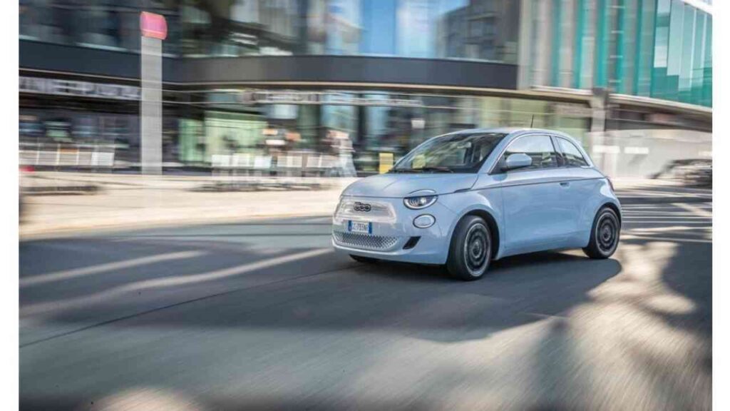 Kompakter, blauer Kleinwagen fährt dynamisch durch urbane Straßen, umgeben von modernen Gebäuden.