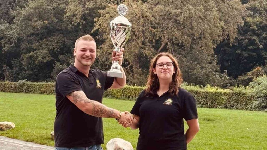 Zwei Personen im Freien schütteln sich die Hand, eine hält einen Pokal hoch, lächelnd vor grüner Kulisse.