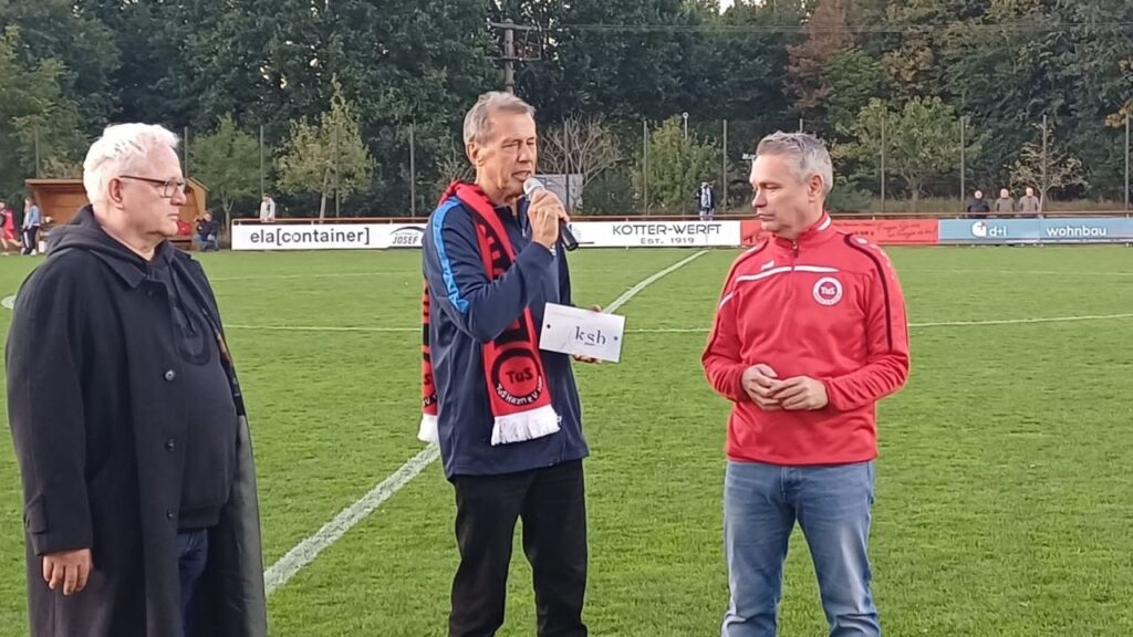 Drei Männer stehen auf einem Fußballplatz, einer hält ein Mikrofon und spricht, zwei tragen Vereinskleidung.