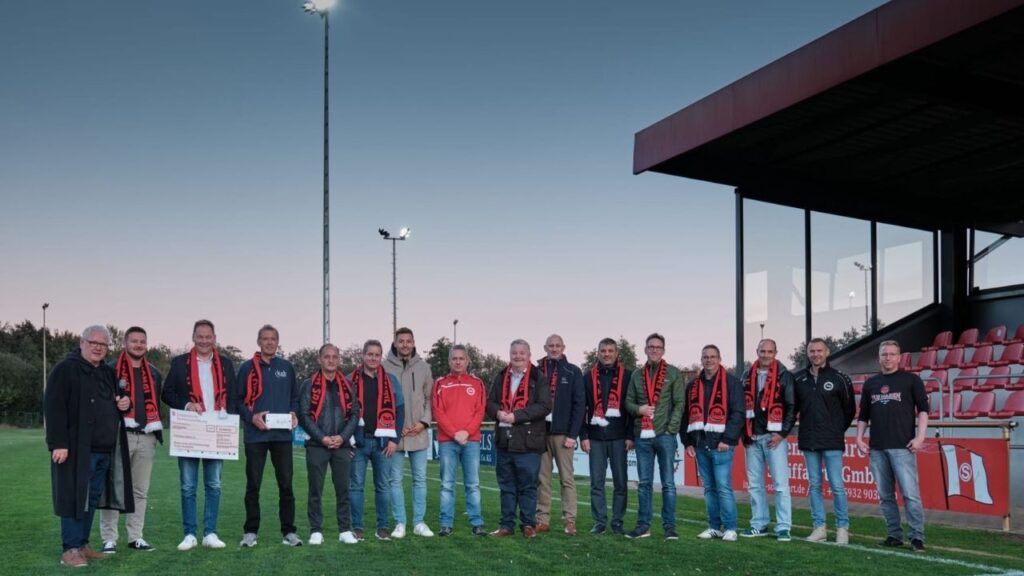 Gruppe von Personen bei einer Fußballveranstaltung im Stadion, einige mit Schals und großen Scheck in der Hand bei Abenddämmerung.