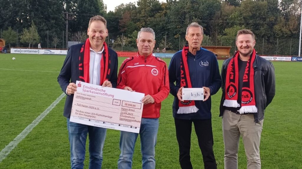 Personen überreichen einen symbolischen Scheck auf einem Fußballfeld, Flutlichtanlage großzügig gefördert.