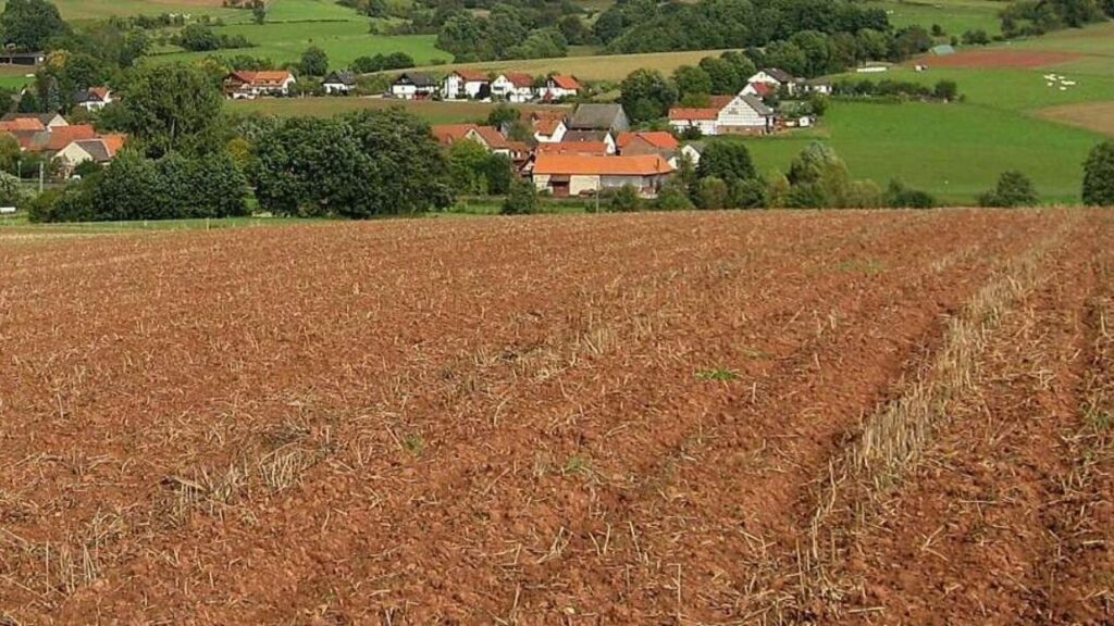Landschaft mit Acker und Dorf im Hintergrund, umgeben von grünen Hügeln und Bäumen unter blauem Himmel.