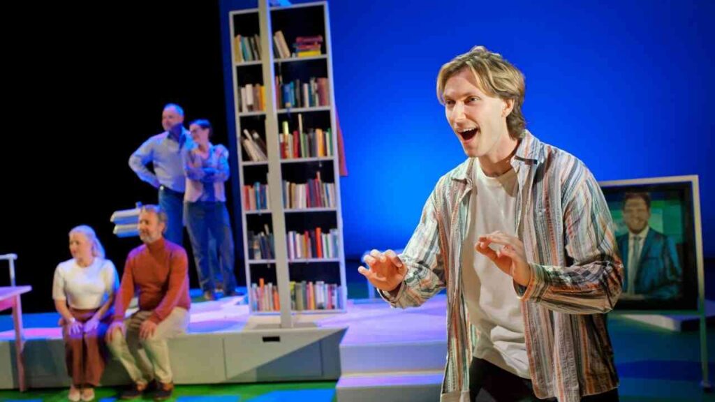 Theaterszene mit Schauspielern vor einem Bücherregal, ein Mann im Vordergrund gestikuliert lebhaft.