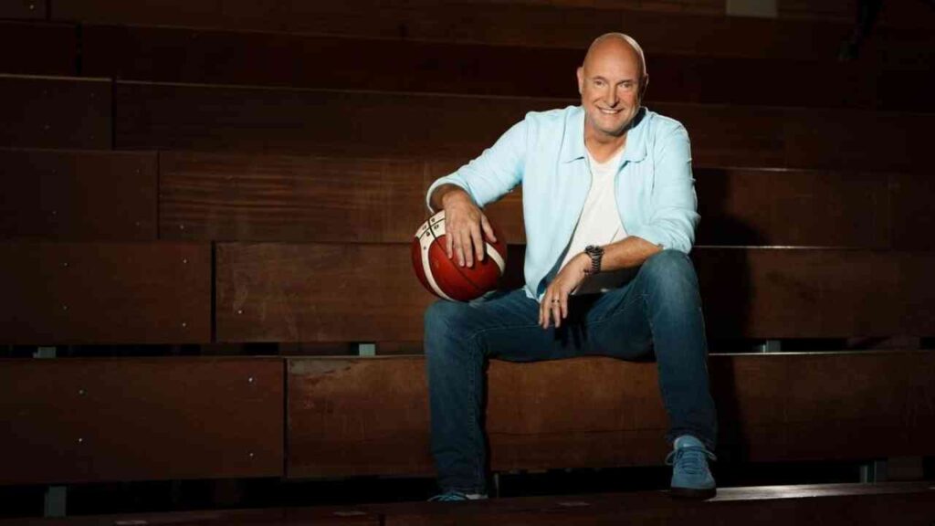 Mann in Freizeitkleidung sitzt mit Basketball auf Tribüne, lächelt in Kamera.