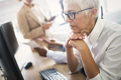 Ältere Frau arbeitet konzentriert am Computer im Büro, im Hintergrund Kolleg*innen in Besprechung.