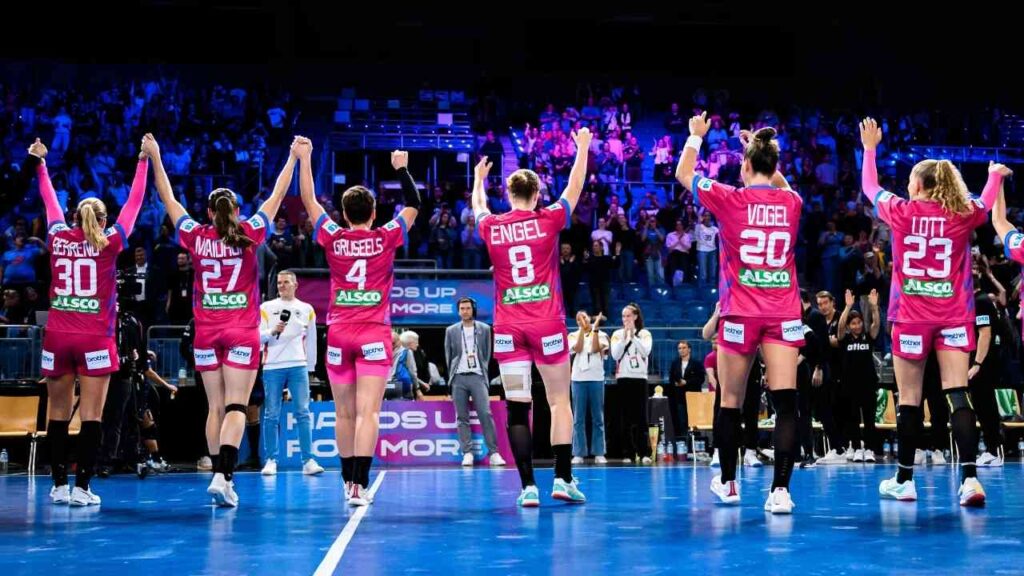 Handballspielerin-Team feiert auf dem Spielfeld mit jubelndem Publikum, Spieler in pinken Trikots heben die Hände.