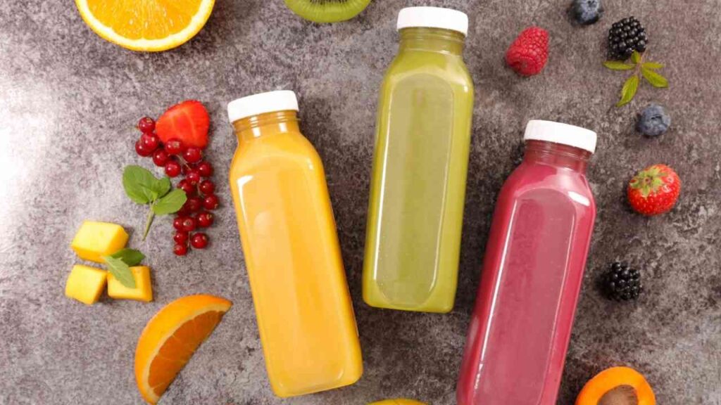 Drei bunte Smoothie-Flaschen aus Mango, Kiwi und Beeren, umgeben von frischen Früchten auf grauem Hintergrund.