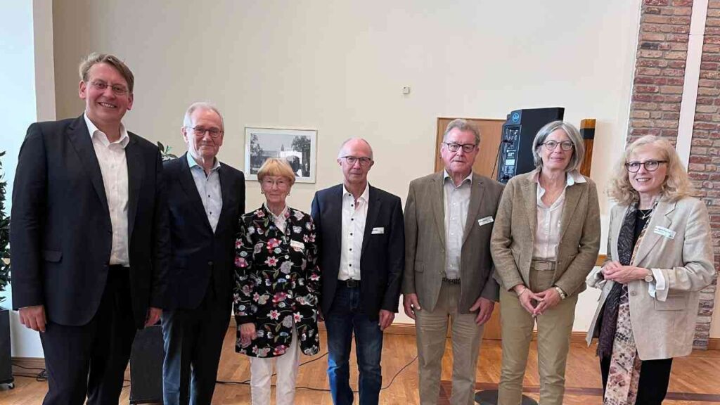 Sieben Personen in formeller Kleidung stehen lächelnd in einem Veranstaltungsraum vor einer Wand und einem Kunstwerk.