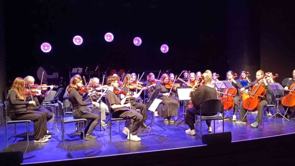 Jugendorchester spielt auf der Bühne klassische Musik, Geige und Cello im Vordergrund, lila Spotlichter im Hintergrund.