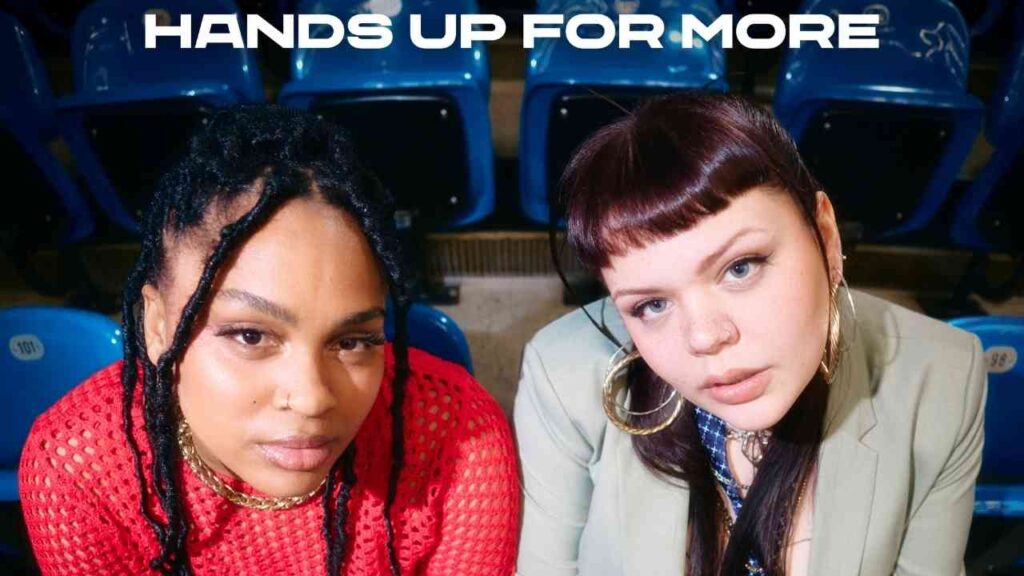 Zwei Frauen in modischer Kleidung in einer Arena, mit Hands Up for More-Text im Hintergrund, Nahaufnahme.