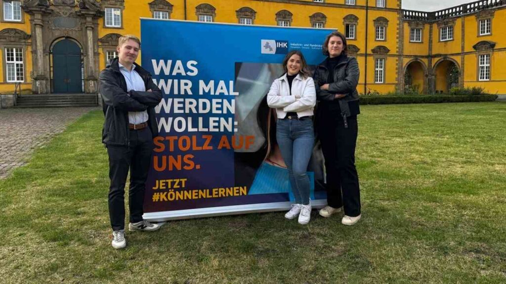 Drei Personen posieren vor IHK-Poster mit Stolz auf uns-Slogan in historischem Gebäude-Hintergrund.