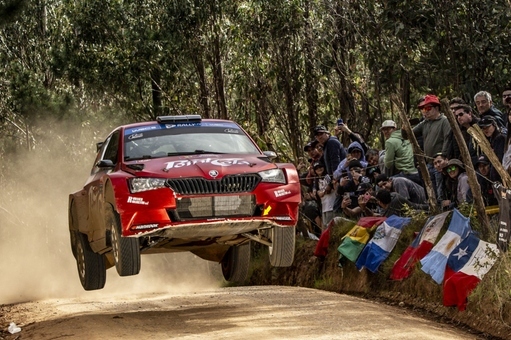 Rallyeauto springt über staubige Strecke, umgeben von Zuschauern und Fahnen, inmitten eines Waldes.