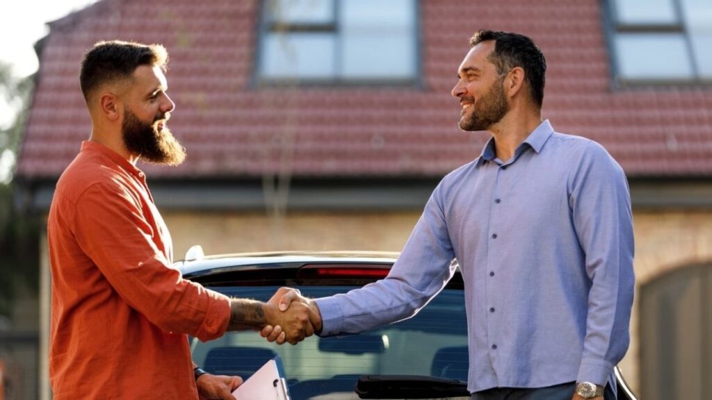 Zwei Männer schütteln sich neben einem Auto die Hand und besiegeln ein Geschäft vor einem Haus.