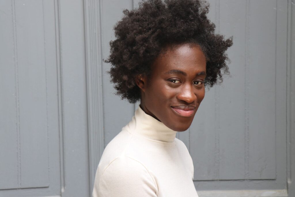 Person lächelt vor grauer Tür, trägt Rollkragenpullover, hat lockiges Haar – entspannter, urbaner Look.