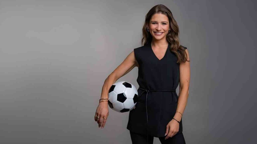 Frau in schwarzem Outfit lächelt mit Fußball im Arm vor grauem Hintergrund.