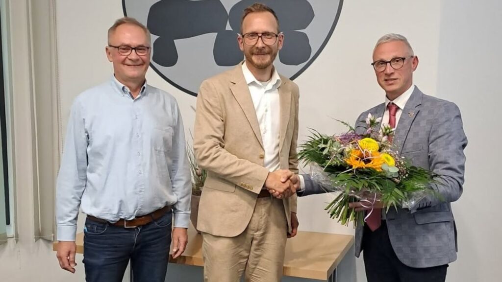 Drei Männer bei einer Gratulation, ein Mann überreicht einen Blumenstrauß und alle lächeln vor einem Firmenlogo.