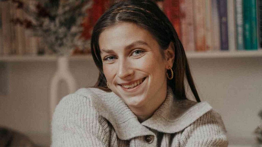 Junge Frau lächelnd vor einem Bücherregal, trägt einen grauen Pulli und Ohrringe. Gemütliche und freundliche Atmosphäre.