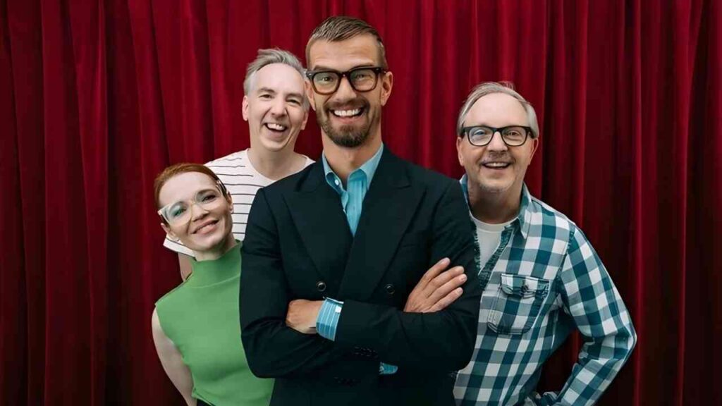 Vier fröhliche Personen lächeln vor rotem Vorhang, eine Person trägt Anzug, die anderen Freizeitkleidung.