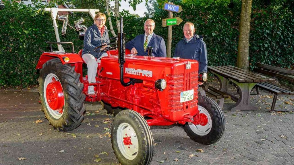 Personen posieren mit einem roten Traktor, umgeben von Bäumen und Schildern im Freien.