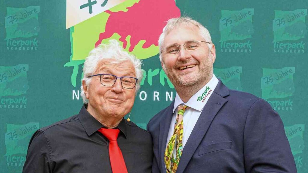 Zwei Männer lächeln vor einem Tierpark Nordhorn-Logo zum 75-jährigen Jubiläum der Einrichtung in festlicher Kleidung.