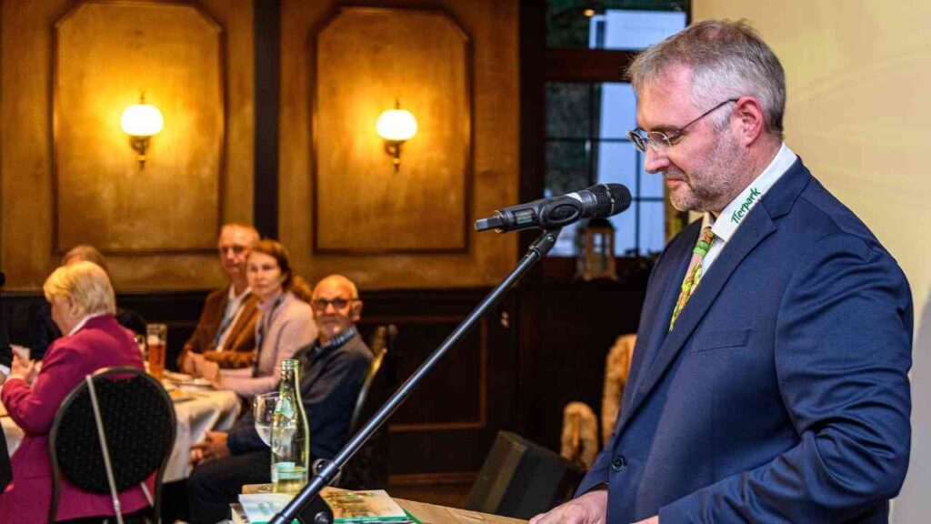 Mann hält Rede bei einer Veranstaltung, Publikum im Hintergrund aufmerksam zuhörend.