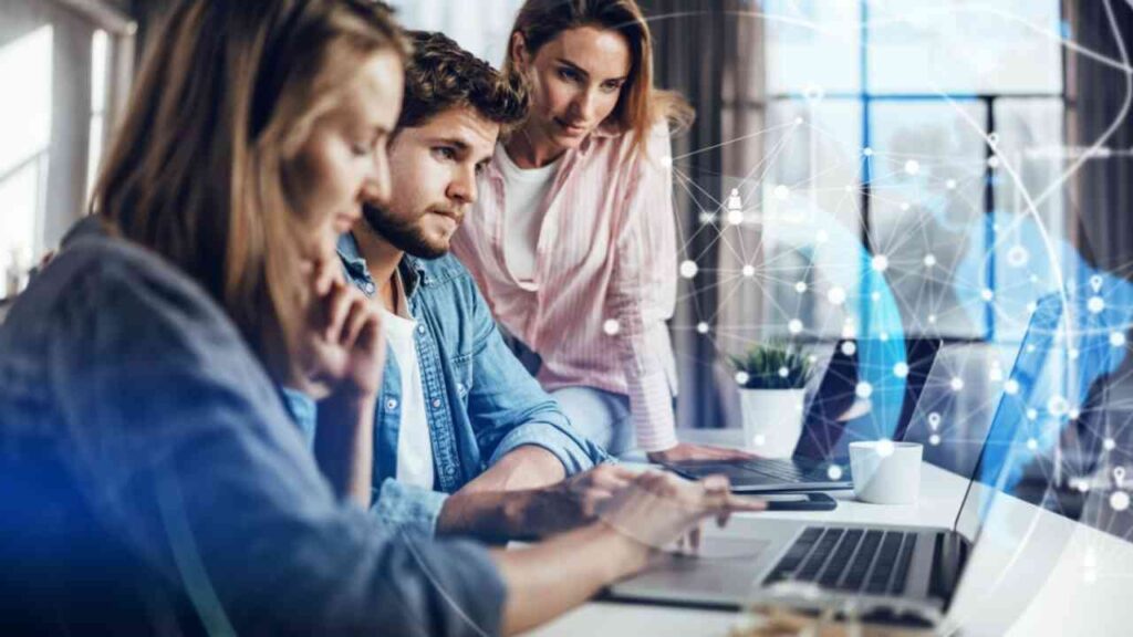 Drei konzentrierte Personen arbeiten gemeinsam an einem Laptop, vernetzte Daten als Overlay, modernes Büro.