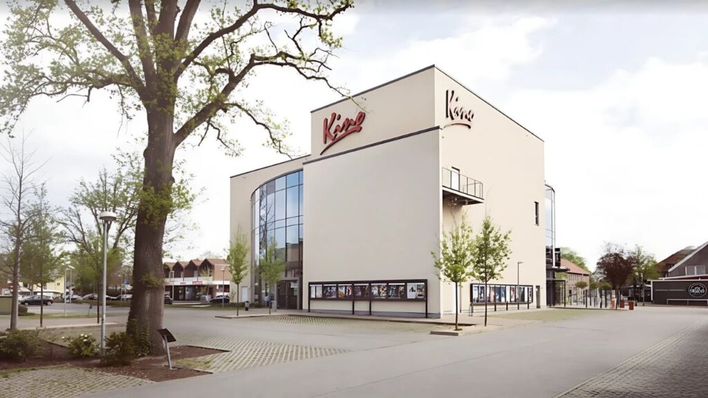 Modernes Kino mit Glasfassade und schlichter Architektur, umgeben von Bäumen, an einem sonnigen Tag.