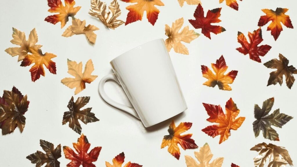 Weiße Tasse auf hellem Hintergrund, umgeben von bunten Herbstblättern. Perfekt für herbstliche Stimmung!