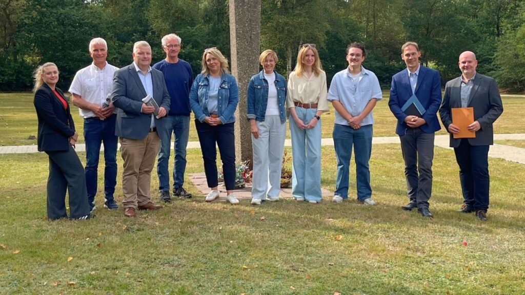 Gruppe von zehn Personen steht im Freien vor einem Denkmal auf einer Wiese bei sonnigem Wetter.