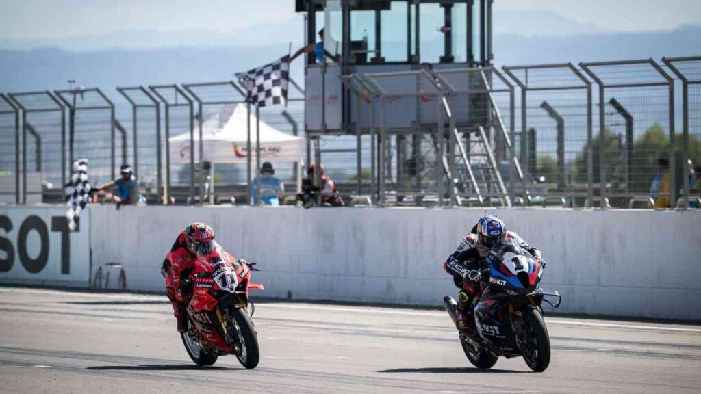 Zwei Motorräder im schnellen Rennen auf einer Rennstrecke, Ziel mit karierten Fahnen, Zuschauer und Zieldurchgang.