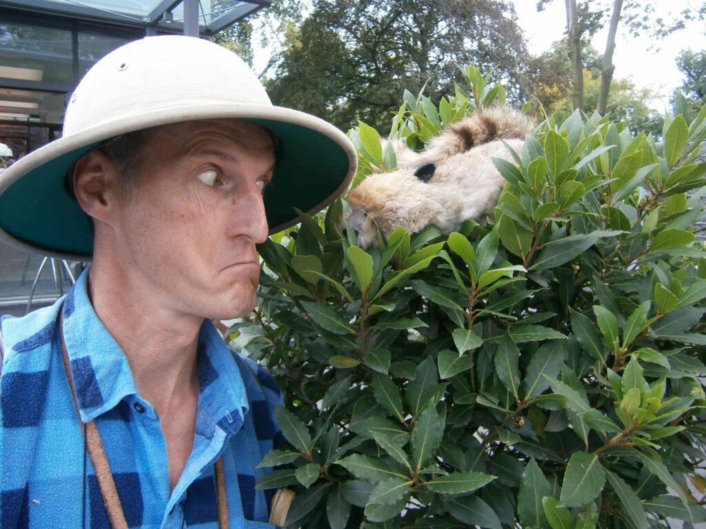 Ein Mann mit Tropenhelm schaut überraschend in einen Busch, hinter dem ein Tierkostüm hervorschaut. Outdoor-Humor.