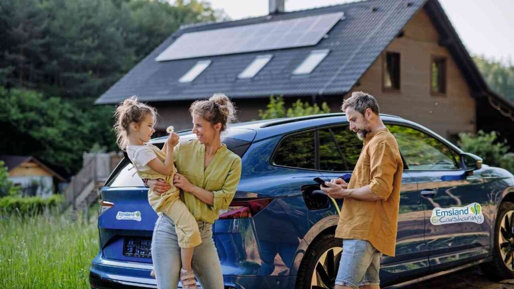 Familie vor einem Emsland Carsharing-Auto bei einem Holzhaus mit Solarpanelen im Grünen. Nachhaltigkeit und Mobilität.