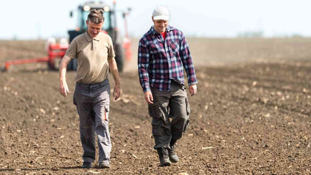 Landwirte gehen über Acker, Traktor im Hintergrund, Vorbereitung für die Aussaat. Landwirtschaft und Zusammenarbeit.