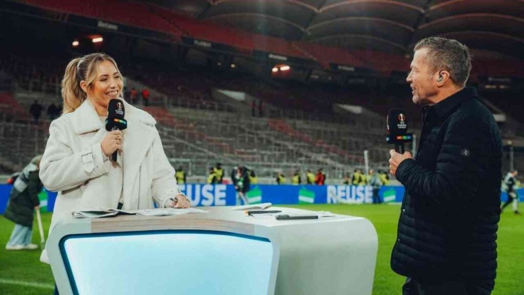 Zwei Sportkommentatoren sprechen im Stadion, Mikrofone in der Hand, beleuchtetes Spielfeld im Hintergrund.