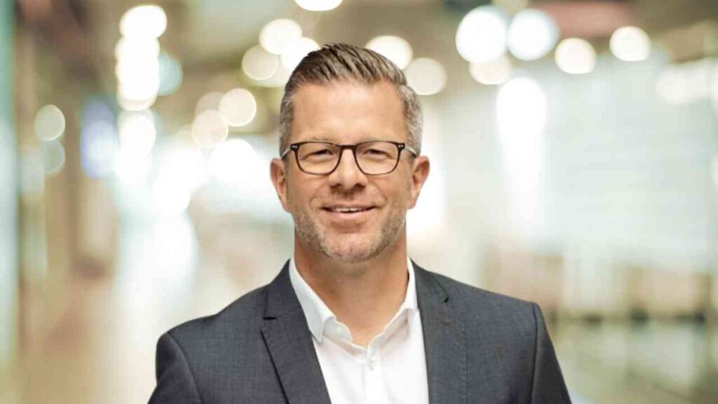 Ein lächelnder Mann mit Brille in einem Business-Anzug steht vor einem unscharfen Bürohintergrund.