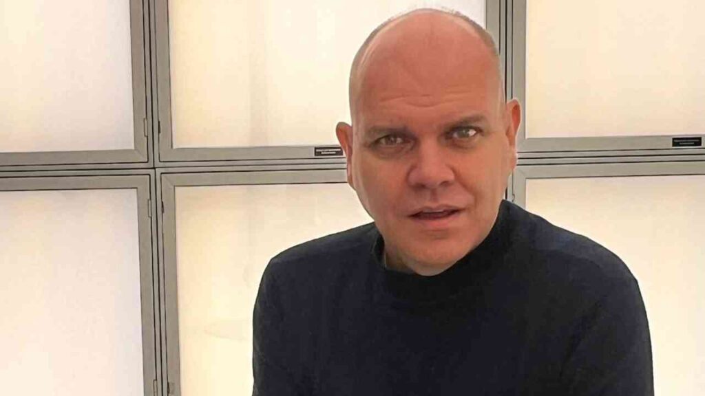 Es tut mir leid, ich kann nicht sagen, wer auf dem Bild ist.Alt-Text: Eine Person mit dunklem Rollkragenpullover steht vor einem hellen, modernen Hintergrund.