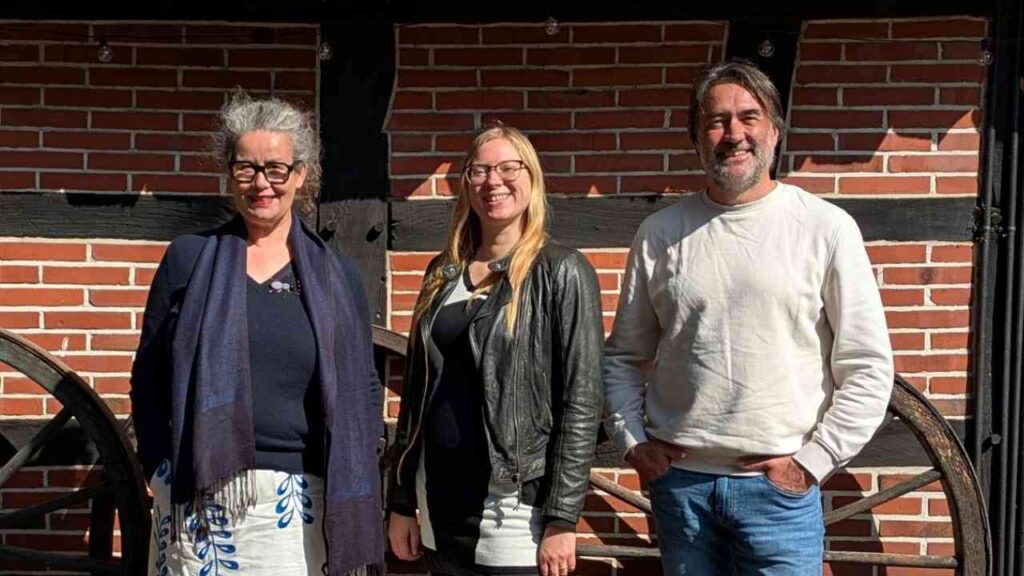 Drei Personen posieren vor einer Ziegelwand mit Holzbalken, lächelnd, bei strahlendem Sonnenschein.