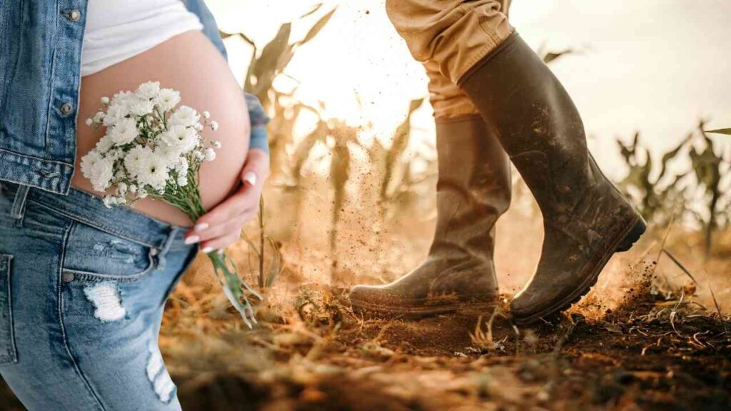 Schwangere Frau in Jeans hält Blumenstrauß vor Babybauch, neben ihr arbeitet jemand in einem Feld mit Gummistiefeln.