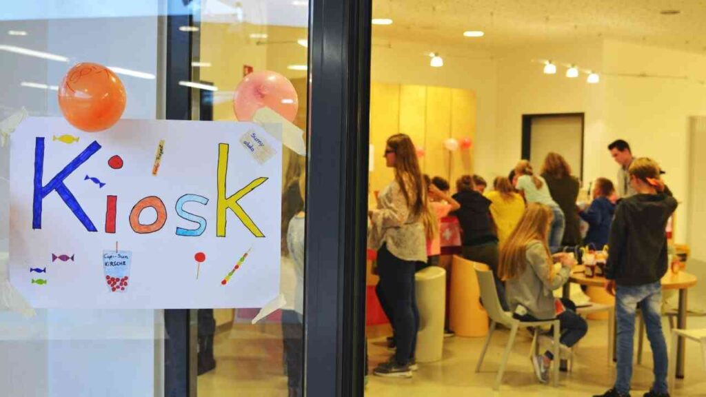 Kiosk-Event mit Kindern und Luftballons in einem Raum, buntes Plakat an der Glastür, fröhliche Atmosphäre.