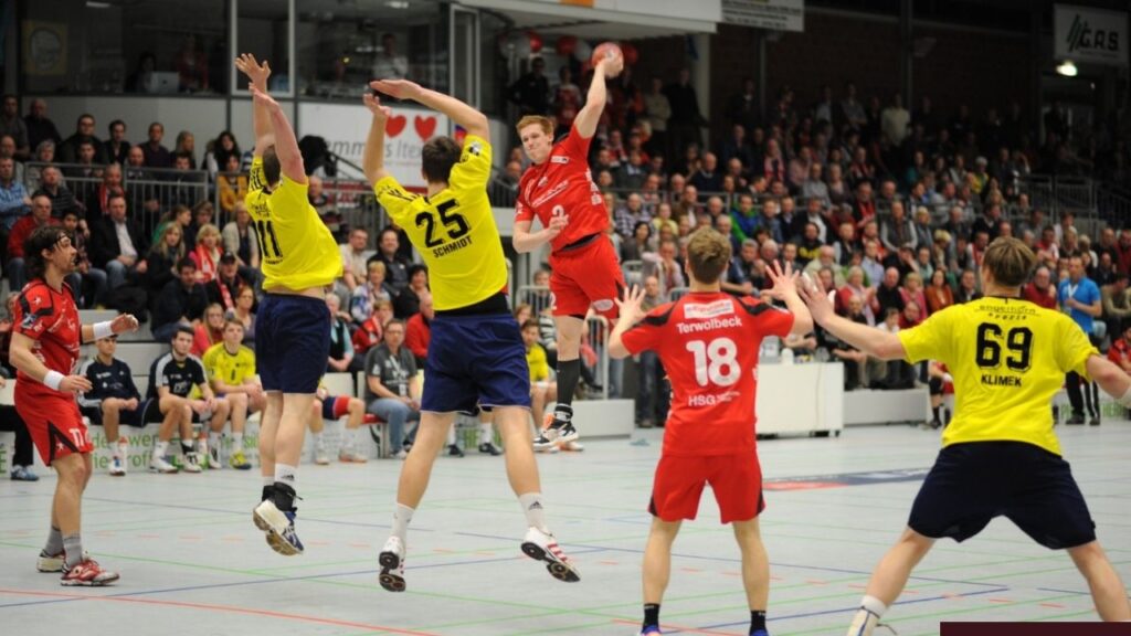 Handballspiel: Spieler im roten Trikot springt zum Wurf, Gegenspieler in gelben Trikots blocken, volle Arena im Hintergrund.