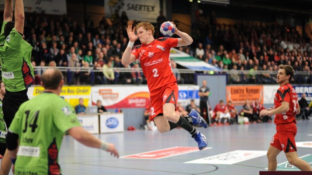 Handballspiel: Spieler in rotem Trikot springt mit Ball, Publikum im Hintergrund, spannende Spielsituation.